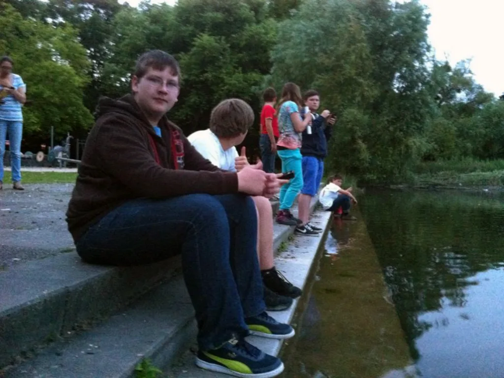 Jugendliche sitzen auf einer Steinstufe am Weiherufer, dahinter weitere Gruppen.